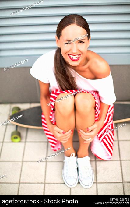 Happy hipster lady sitting on longboard with one eye closed. Toothy smiling girl in T-shirt and skirt holding her slim and slender legs