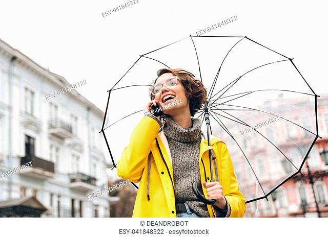 Shot of young woman in yellow raincoat and glasses talking on mobile phone, having pleasant conversation under umbrella