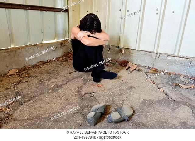 A sad woman is sitting alone on the cold concrete behind a shed