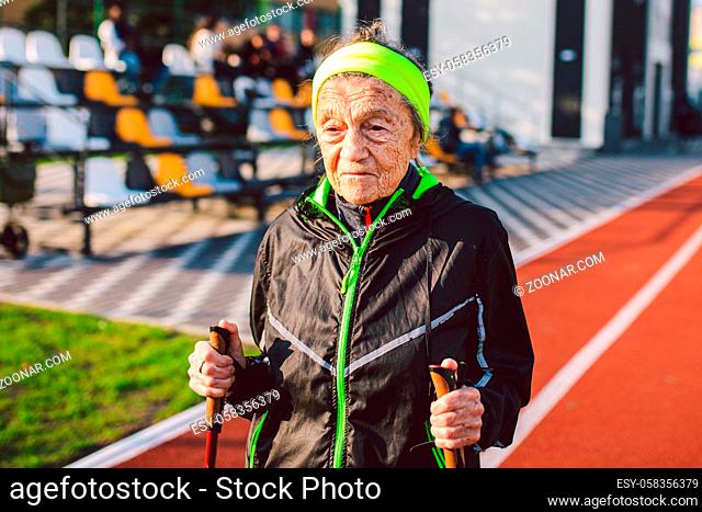 Active elderly Caucasian elderly women of 90 years practice Nordic walking with ski poles on a track with a red rubber coating. Active holidays