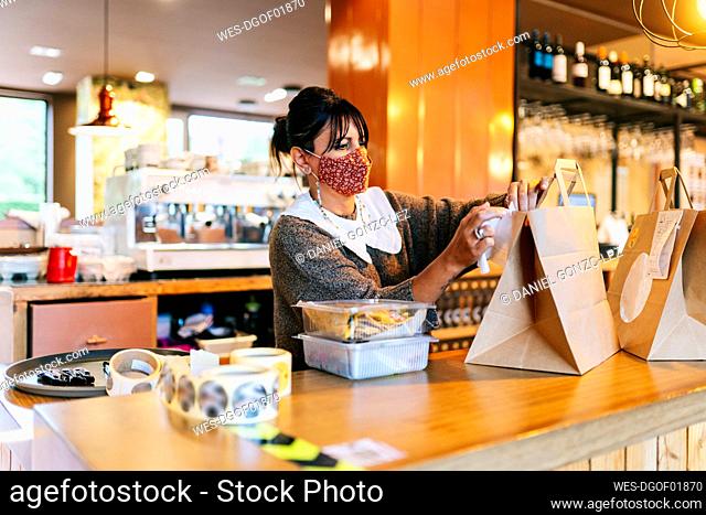 Female owner in mask attaching receipt on brown paper bag on counter at restaurant