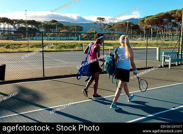 Full length of multiracial female tennis players walking with rackets and bags at court on sunny day