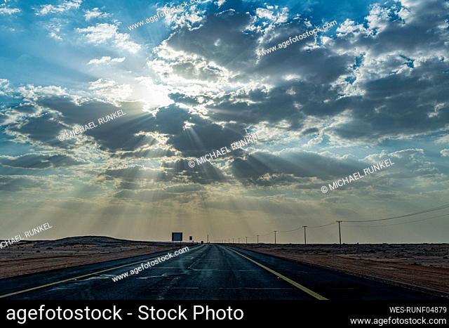 Saudi Arabia, Al-Ula, Setting sun piercing clouds over empty highway