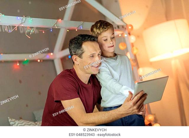 Show it to me. Smiling man holding modern device in right hand and sitting near his son while watching video together