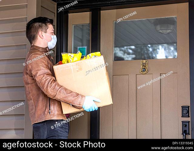 A young man standing with a box of groceries at the door of a house during the Covid-19 World Pandemic; Edmonton, Alberta, Canada