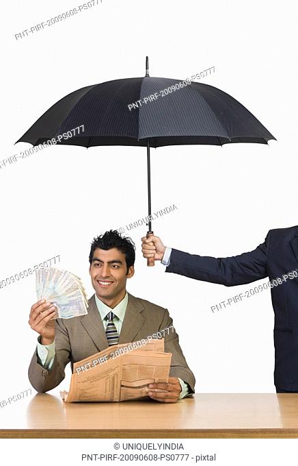Businessman holding a newspaper and currency notes under umbrella