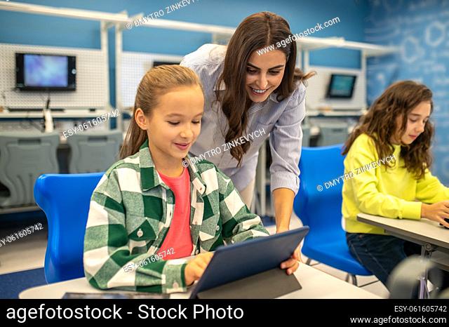 Lesson, pleasure. Smiling young woman looking into tablet of contented girl sitting at desk studying during lesson
