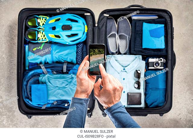 Overhead view of man's hands using smartphone touchscreen above packed suitcase with blue bike helmet, backpack, retro camera and shirt