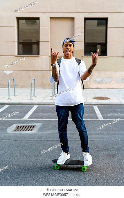 Tattooed young man standing on skateboard sticking out tongue and showing Rock And Roll Sign