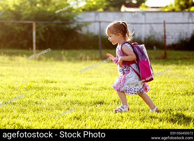 The cute little girl with schoolbag is walking on grass