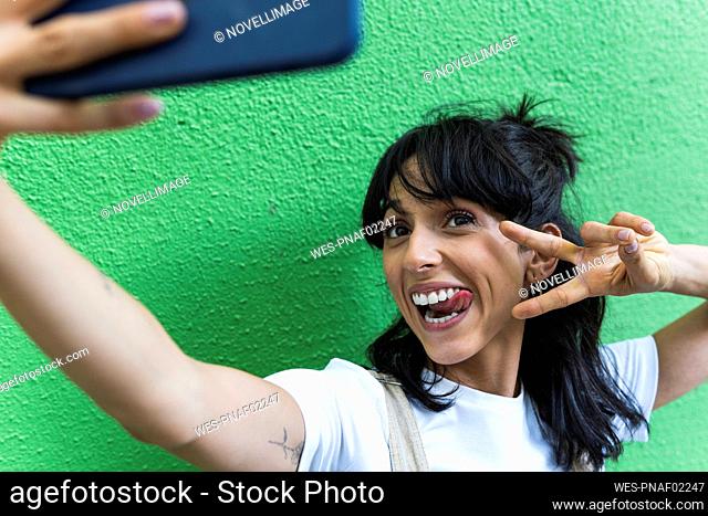 Cheerful woman showing peace sign and sticking out tongue while taking selfie