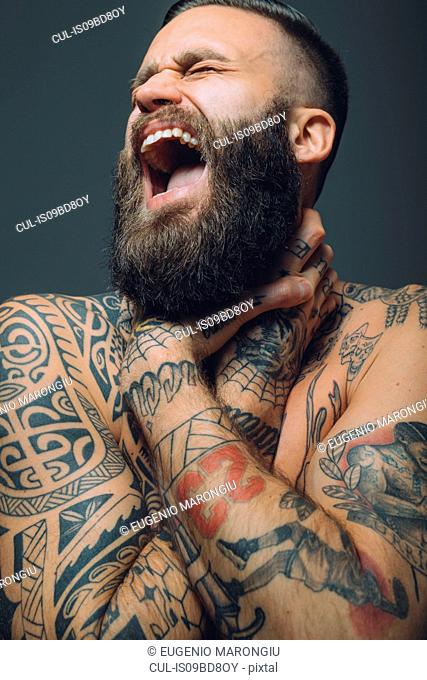 Portrait of young man with beard, covered in tattoos, hands around throat, screaming