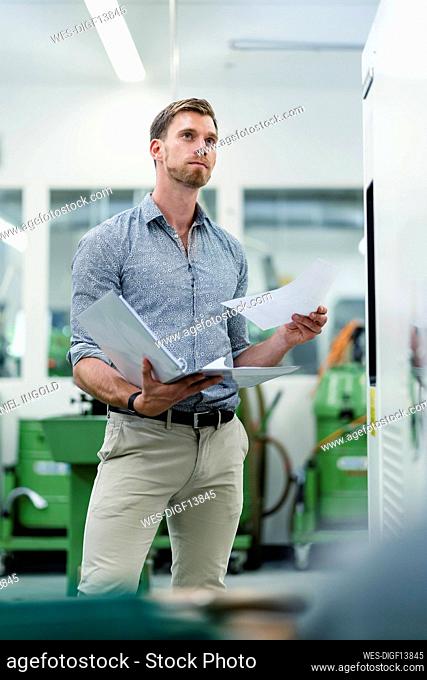 Businessman with file document day dreaming while standing by machine in factory
