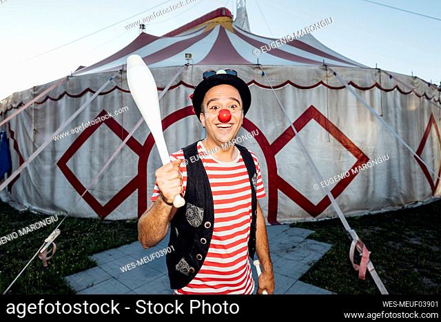 Smiling male clown holding juggling pin while standing in front of circus tent