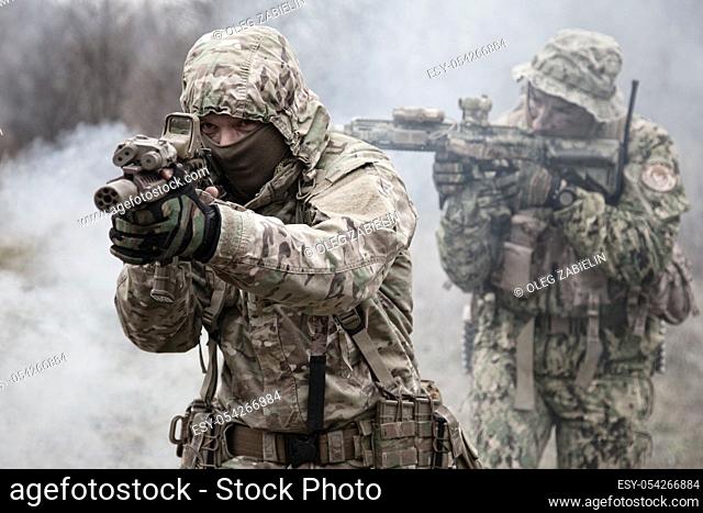 Two soldiers in camouflage uniform, wearing military ammunition, aiming service rifles, covering each other, shooting in competitors