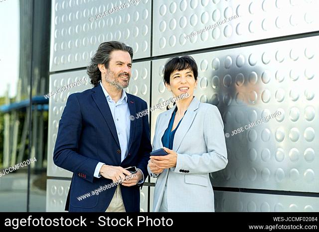 Thoughtful mature entrepreneurs with mobile phone standing by silver colored wall
