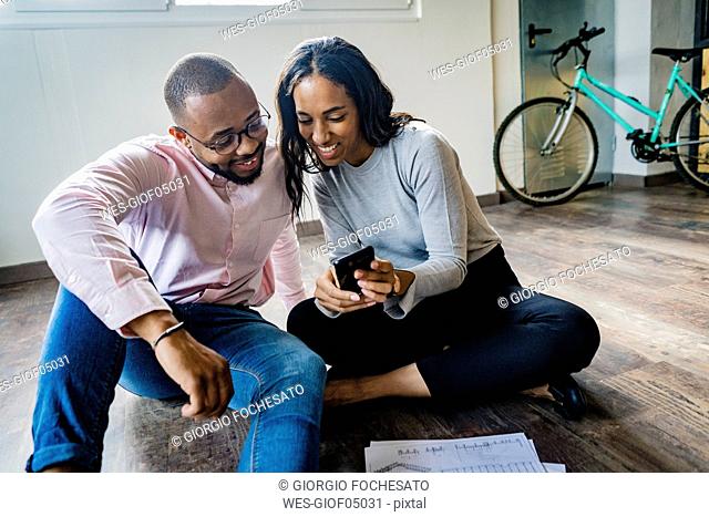 Smiling businessman and businesswoman sitting on the floor with cell phone and documents