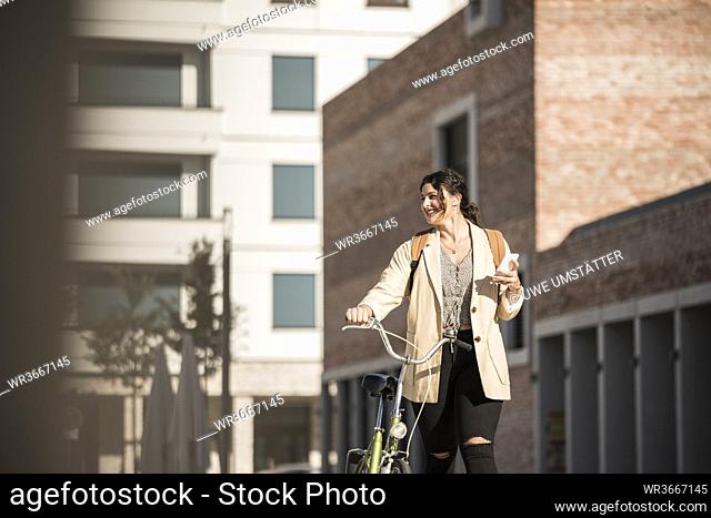 Smiling female student looking away while walking with bicycle against buildings