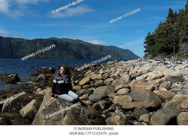 Woman sitting on rocks at coast, Norris Point, Bonne Bay, Gros Morne National Park, Newfoundland And Labrador, Canada