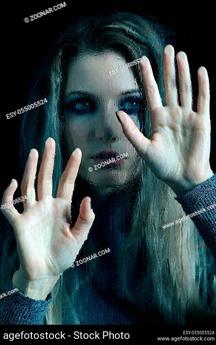 Young woman behind the window glass with raindrops. Female in gray sweater looking through the glass on black background