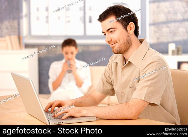 Young man using laptop, browsing internet at home, woman sitting in background, drinking tea