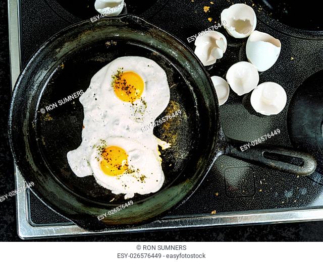 Photo of two eggs frying in a cast iron skillet on top of a stove with egg shells to the side