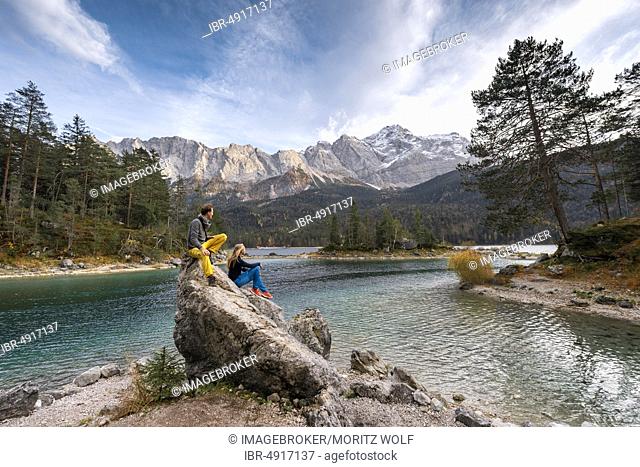 Two hikers sitting on a rock on the shore, view into the distance, Eibsee lake in front of Zugspitzmassiv with Zugspitze, Wetterstein range, near Grainau