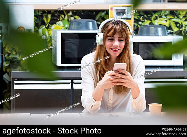 Smiling businesswoman wearing headphones surfing net through smart phone in cafeteria