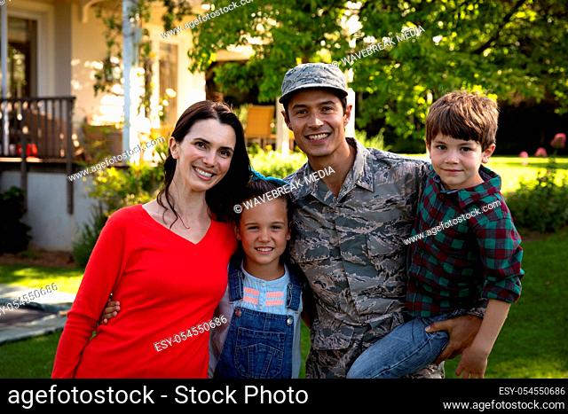 Portrait of a young adult mixed race male soldier in the garden outside his home, standing with his arms around his Caucasian wife and young daughter