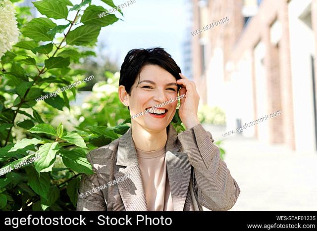 Cheerful businesswoman touching hair by plant on sunny day