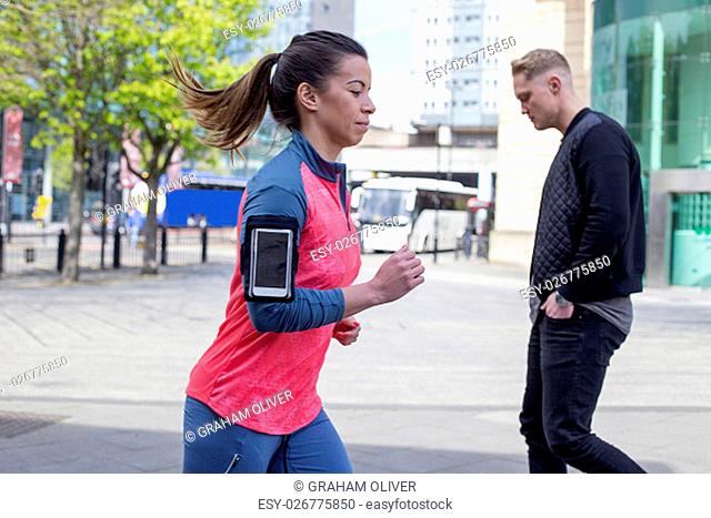 Woman running in the city. She has a smartphone in her arm strap. There is a man walking behind her