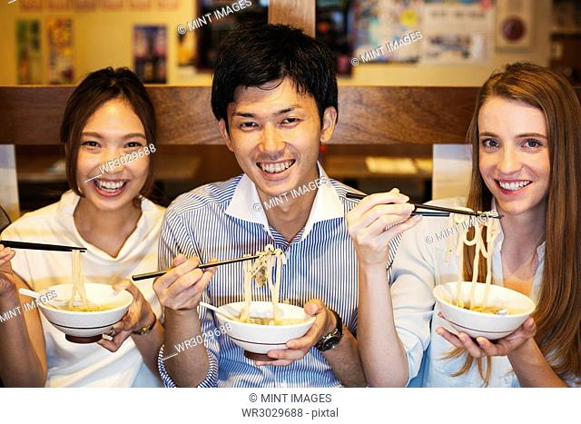 Three smiling people sitting sidy by side at a table in a restaurant, eating from bowls using chopsticks