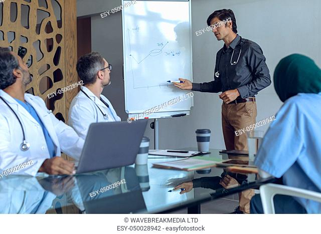 Front view of Caucasian male doctor explaining graph on flip chart in meeting with diverse medical team at hospital. Coffee cup, medical folders, clipboard