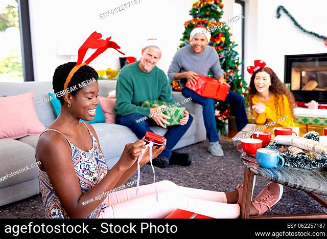 Image of happy african american woman opening gift, celebrating christmas at home with friends