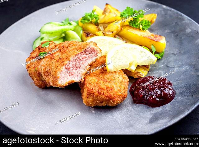 Traditional deep fried veal steak with fried potatoes and cucumber salad offered as closeup on a modern design plate