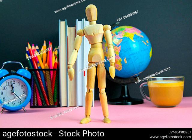 Figure of a wooden man is in the foreground and behind him are a stack of books, a globe, a clock and a glass of pencils
