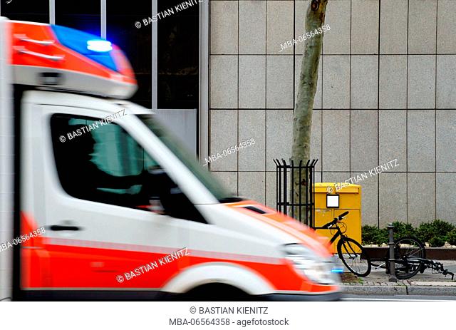 Photography of parked bicycles and a motor scooter in front of a mailbox with an ambulance with blue light driving past