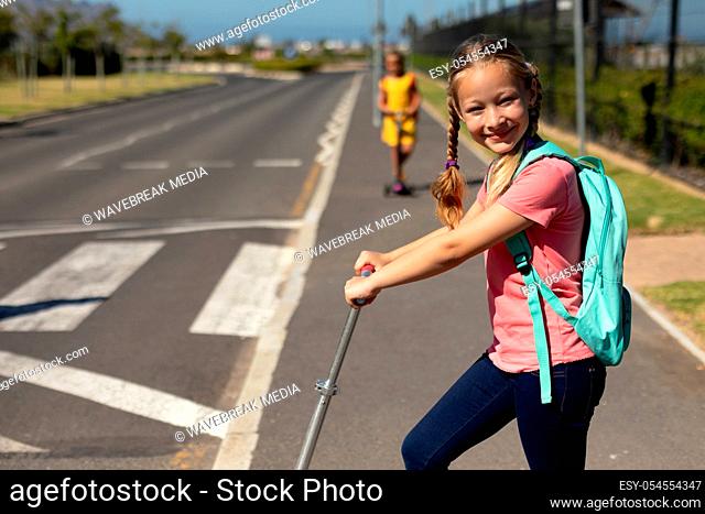 Side view of a Caucasian schoolgirl with plaits wearing a pink t shirt and a rucksack standing on a push scooter on the pavement
