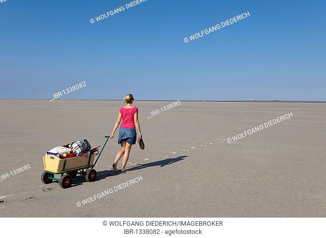 Young woman, 20-25 years, pulling a wagon with luggage across the sandy beach of St Peter Ording, North Sea, North Friesland, Schleswig-Holstein