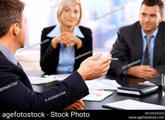 Hand in closeup holding pen at business meeting, coworkers listening in the background. Focus on hand
