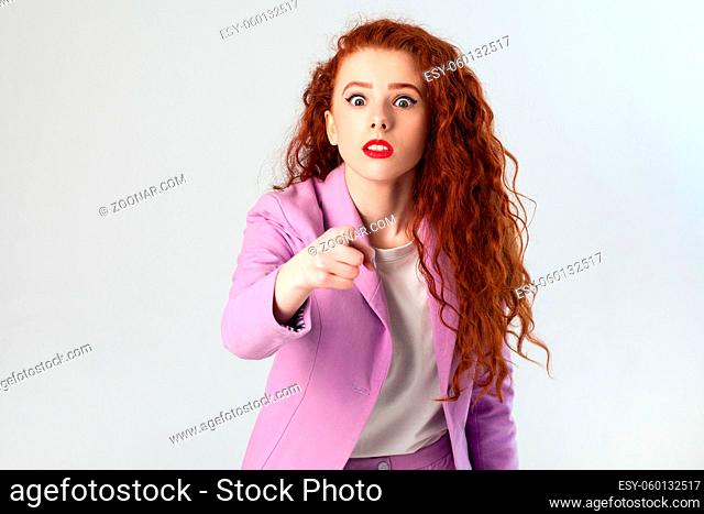 Portrait of shocked beautiful business woman with red - brown hair and makeup in pink suit. pointing and looking at camera, studio shot on gray background