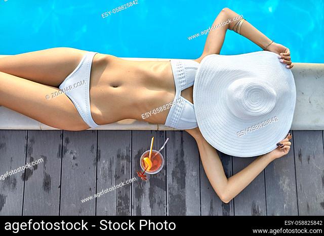 Trendy girl in a white swimsuit lies on the pool's edge outdoors. She holds a big hat which closes her face. Next to her there is an orange cocktail