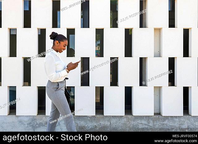 Smiling businesswoman using mobile phone while walking by block wall