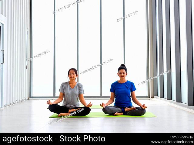 Two Asian women sporty attractive people practicing yoga lesson together, working out at the fitness GYM, Young and senior female exercising do yoga in yoga...