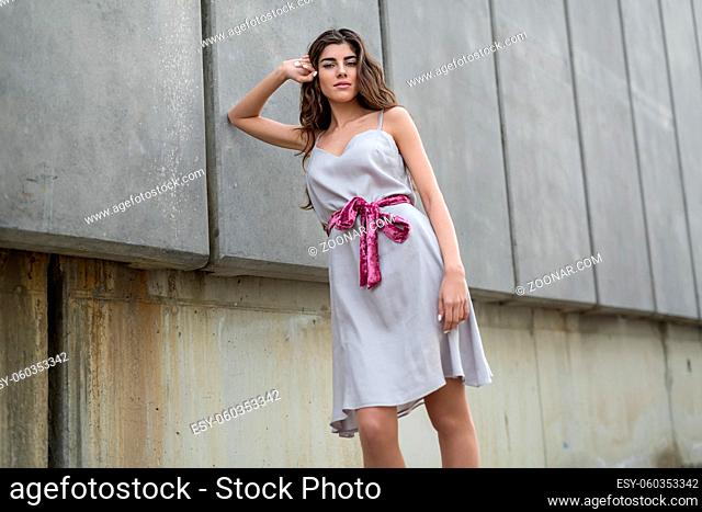 Pretty brunette with beautiful hair stands on the background of the concrete wall. She holds her right elbow on the wall and the right hand on the head