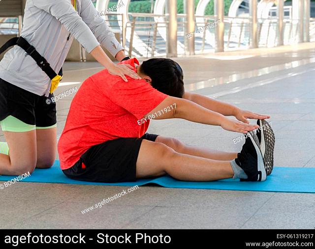 Instructor supporting overweight woman while doing stretching exercises outdoors. Plus size young woman sitting and touching her toes on yoga mat