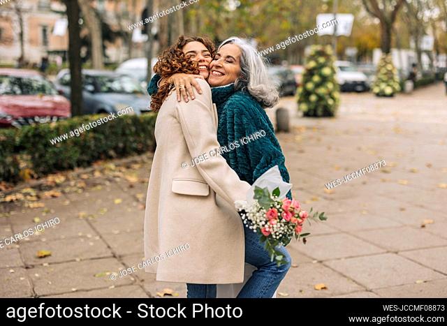 Cheerful young woman with bouquet of flowers hugging grandmother at footpath