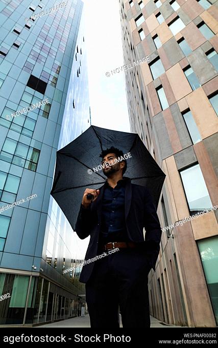 Entrepreneur with hand in pocket holding umbrella against building in city
