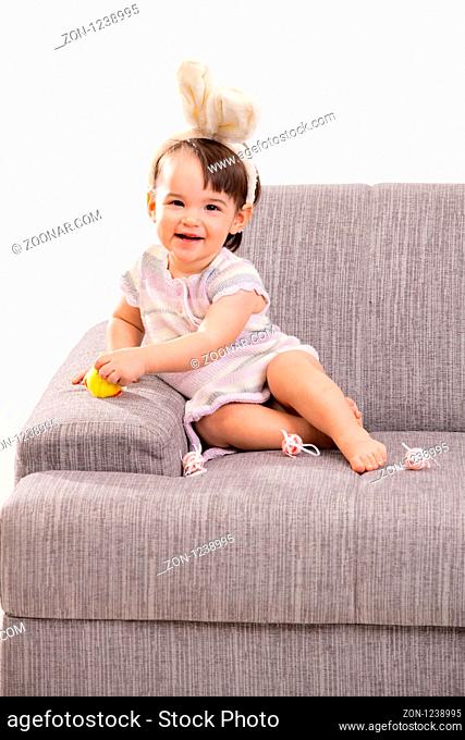 Baby girl in easter bunny costume, sitting on grey couch playing with toy chicken and easter eggs, laughing. Isolated on white background