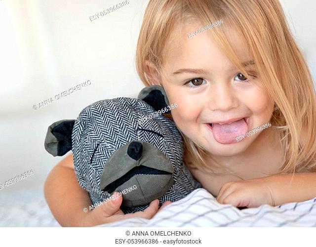 Closeup portrait of a cute baby boy making faces, showing the tongue, having fun at home with his soft toy, happy healthy carefree childhood